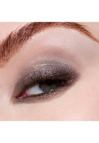 Nahaufnahme eines Auges mit rauchigem grauem Lidschatten und dunklem Eyeliner. Die Wimpern sind definiert. Die Haut ist glatt mit einem hellen Pfirsichton. Darüber befindet sich eine braune Augenbraue.