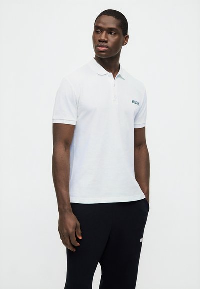 Άνδρας που φοράει λευκό κοντομάνικο πόλο με το κείμενο "Moschino" και μαύρο παντελόνι, στέκεται μπροστά σε απλό λευκό φόντο.