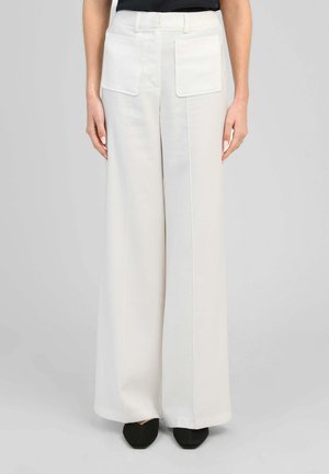 Femme portant un pantalon blanc taille haute à jambes larges avec de grandes poches plaquées devant et des chaussures noires pointues, debout devant un fond uni.