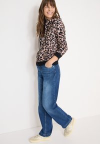 Reißverschluss-Hoodie mit Leopardenmuster in Pink und Schwarz, kombiniert mit blauen Jeans mit geradem Bein und hellen Sneakers mit beigen und weißen Akzenten.