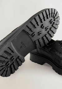 Scarpe in pelle nera con suola in gomma testurizzata caratterizzata da scanalature profonde e forma arrotondata. I lacci e le cuciture sono visibili sulla parte superiore.