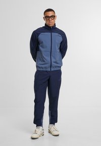 Jūras spēku un gaiši zila sporta uzvalks ar aizdari žaketi un elastīgu jostasvietu biksēm. Iezīmēts ar logo uz krūšu daļas un teksturētu neilona audumu.