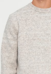 Gros plan sur une personne portant un pull en tricot texturé gris clair avec un col rond.