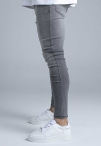 Szürke szűk fazonú farmer fehér platform tornacipővel, amelyek texturált díszítésekkel és fűzőkkel rendelkeznek, oldalnézetből mutatva.