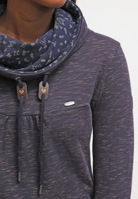 Žena v modrém mikinovém svetru s tečkovaným vzorem, s dvojitou kapucí a květinovou vnitřní vrstvou, tlustými šňůrkami visícími dolů.