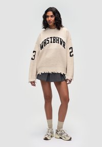 Übergroßer beiger Strickpullover mit "WRSTBHVR" und "23" in Schwarz. Mit einem ausgefransten Saum und weiten Ärmeln. Kombiniert mit einem grauen Rock und Sneakern.