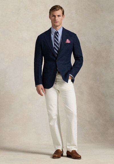 Sötét tengerészkék blézer, csíkos kék-fehér ing, rózsaszín zsebkendő, krémszínű nadrág, barna cipő; testre szabott, strukturált kialakítás.