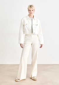 White cropped denim jacket with silver buttons, paired with high-waisted white flared trousers and beige sneakers with white accents.