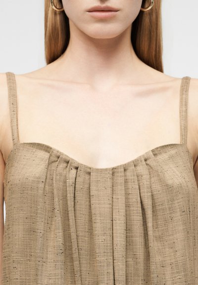 Γυναίκα με αμάνικη μπεζ τοπ με υφή και πτυχωτές λεπτομέρειες, με εκτεθειμένο λαιμό και ώμους, και χρυσούς κρίκους στα αυτιά.