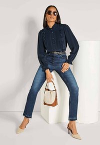 Navy-Bluse mit Rüschendetails, kombiniert mit hochgeschnittenen blauen Jeans und beigen Slingback-Absätzen. Eine Handtasche in Braun und Creme rundet den Look ab.