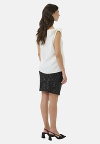 Femme aux longs cheveux bruns portant un haut sans manches blanc avec un nœud à l'épaule, une jupe noire pailletée et des escarpins à talons noires.