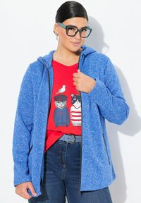 Blå fleecetröja med texturmateriel, framdragkedja och sidofickor. Kombinerad med en röd grafisk t-shirt med kattillustrationer.