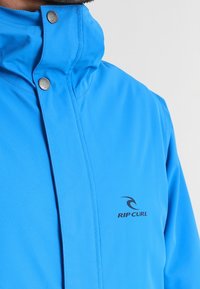 Kirkas sininen vedenpitävä takki, jossa on korkea kaulus, painonapit ja musta "RIP CURL" -logo. Sileä pinta ja kevyt materiaali.