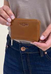 Hnedá kožená peňaženka s embosovaným logom, so zipsom, hladký povrch, držená v ruke, s rustikálnym prešívaním a strieborným ťaháčikom na zips.