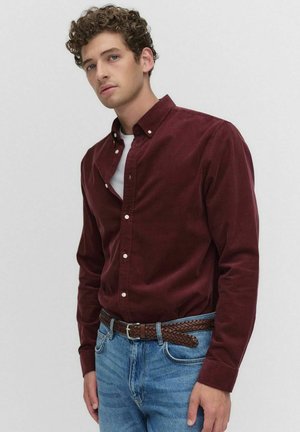 Jonge man in een knopenburgundy shirt over een wit hemd, blauwe spijkerbroek en een gevlochten bruine riem, staand tegen een effen achtergrond.