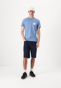 Lys blå bomull t-skjorte med et mønstret brystlomme, sammen med marineblå shorts, hvite sokker og svarte sko. Beige caps fullfører antrekket.