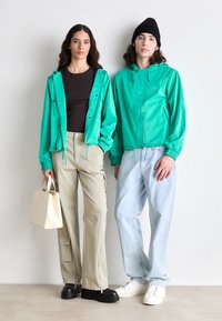 Două persoane poartă jachete verzi strălucitoare, pantaloni cargo bej și blugi bleu deschis. Una dintre ele poartă o geantă mică crem.