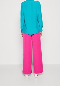 Türkise Bluse mit langen Ärmeln, kombiniert mit weiten rosa Hosen. Glatte Textur, minimales Design und klassischer Kragen. 