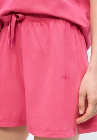 Femme portant un short ample rose avec une taille élastique et un cordon de serrage noué, la main reposant près de l'ourlet, logo "BOSS" en relief visible.