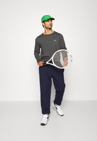 Maglietta grigio a maniche lunghe con un piccolo logo, pantaloni sportivi blu navy, cappellino verde e una racchetta da tennis bianca, completata con sneaker bianche.