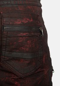 Denim nero con un motivo rosso strappato, dotato di tasca posteriore con zip e tessuto testurizzato. Linee pulite e un aspetto robusto.