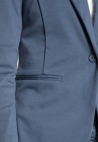 Modrý sako na míru s jedním knoflíkem a klopovým kapsou vpředu, nošený přes bílou košili.