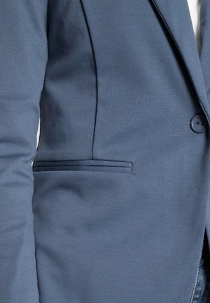 Modrý blejzr s úzkým střihem, hladkým texturovaným materiálem, zapínáním na jeden knoflík a boční kapsou na levé straně.