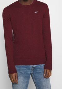 Burgundy stickad tröja med rundad hals, långa ärmar, ribbad mudd och fåll, med en liten vit logotyp på bröstet. Bärs över blå jeans.