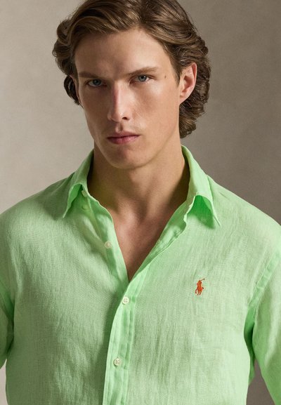 Ανοιχτό πράσινο λινό πουκάμισο με κουμπιά, με ένα μικρό πορτοκαλί κεντημένο λογότυπο στο αριστερό στήθος και χαλαρό γιακά.