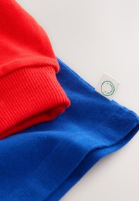 Red long-sleeve cuff folded above a blue garment showing a white tag with a green smiley face on a light background.