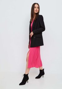 Zwarte op maat gemaakte blazer over een roze mididress met een zijsplit. Zwarte enkelboots met vetersluiting en een smalle hak.