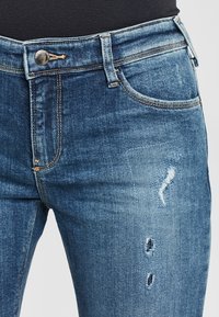 Vybledlé modré džíny s zipem, pěti kapsami a opotřebovanými detaily na přední části. Šité akcenty a kovové zapínání.