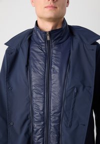 Giacca blu navy con strato interno staccabile in piuma, colletto doppio, chiusura a zip e tasca sul petto con zip. Texture morbida.