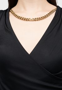Χοντρή χρυσή αλυσίδα με κεντρικό διακοσμητικό «Μ», φορεμένη με μαύρο τοπ με V λαιμόκοψη.
