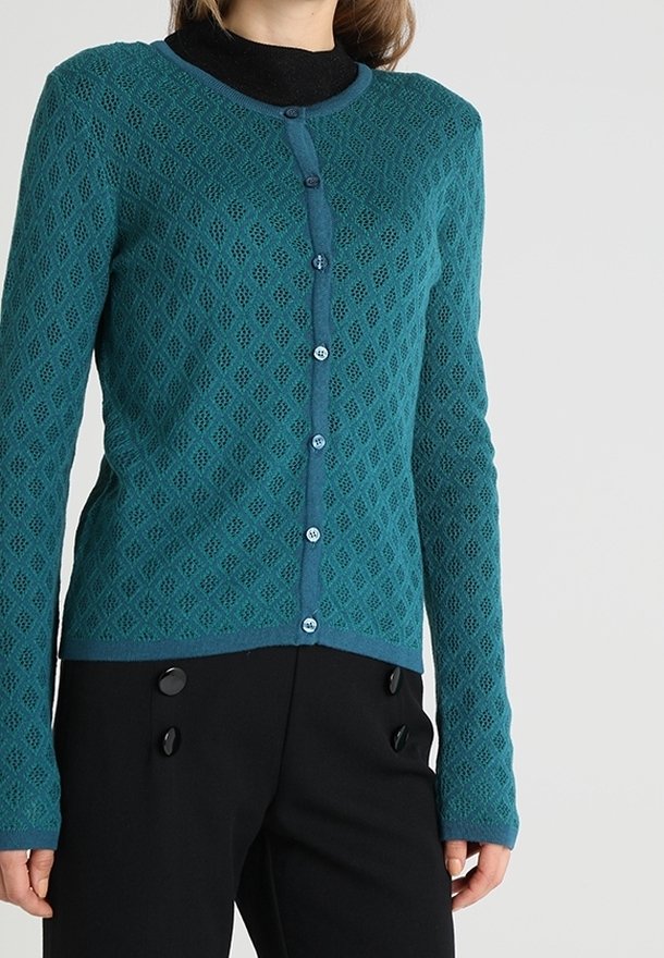 Femme portant un cardigan boutonné à motif losanges couleur sarcelle sur un top noir, associé à un pantalon noir taille haute avec de gros boutons.