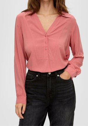 Vrouw draagt een lange mouwen roze blouse met drie knopen en zwarte high-waisted jeans, één hand in de zak tegen een effen achtergrond.