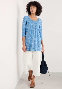 Blauwe tuniek met wit bloemenpatroon, driekwart mouwen, gecombineerd met witte culottes en bruin gespsandalen, terwijl ze een marineblauwe tas draagt.