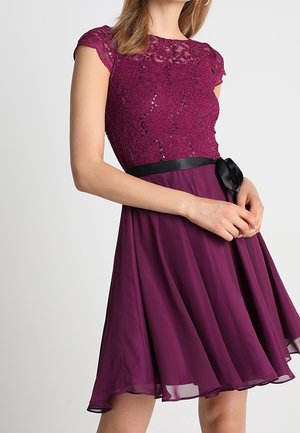 Femme portant une robe de cocktail violette sans manches avec de la dentelle et des paillettes sur le corsage, une ceinture noire et une jupe fluide en chiffon.