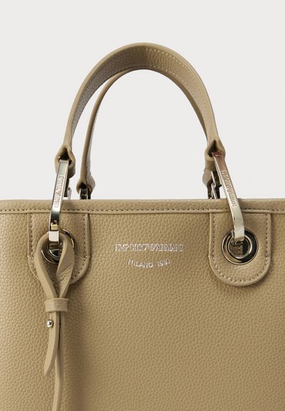 Μπεζ δερμάτινη τσάντα χειρός με υφή φινιρίσματος, ασημένια μεταλλικά στοιχεία, διπλές λαβές και ανάγλυφο λογότυπο. Διαθέτει κρεμαστή δερμάτινη ετικέτα.