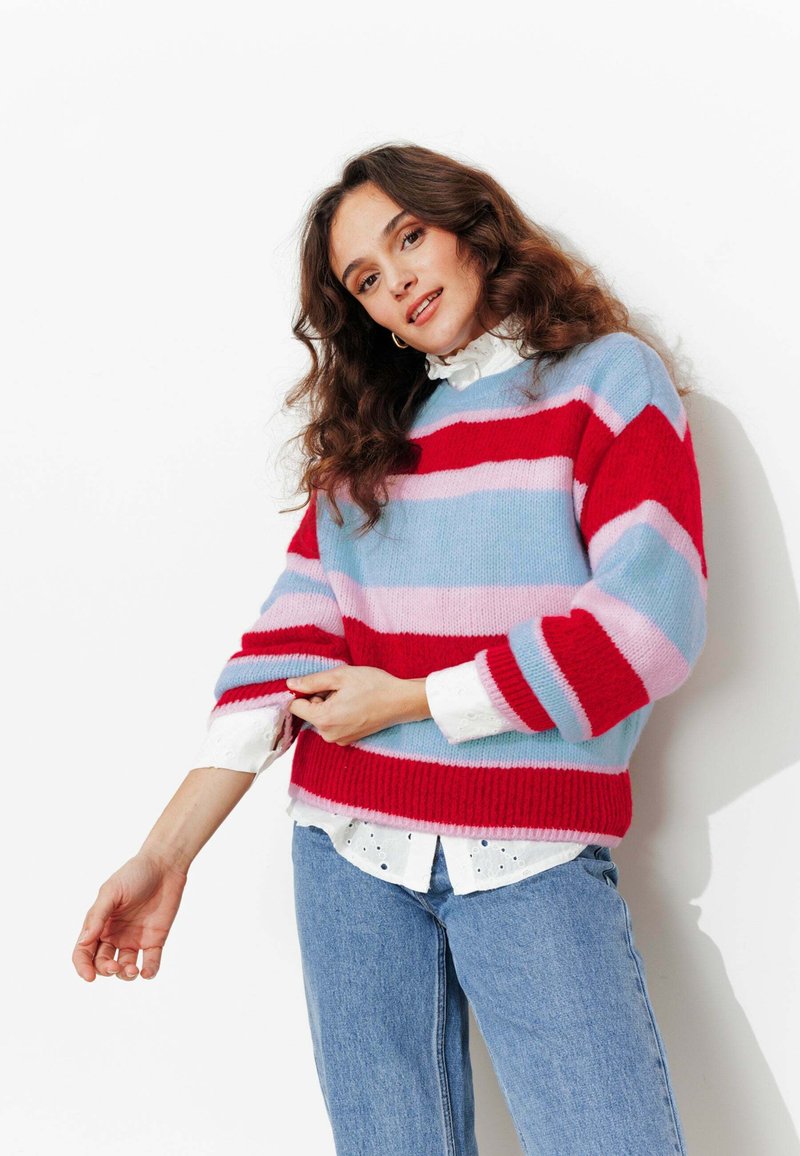 Pull tricoté avec des rayures horizontales rouges, bleues et roses. Présente une coupe décontractée, un col rond et des manches surdimensionnées. Superposé sur un chemisier blanc.