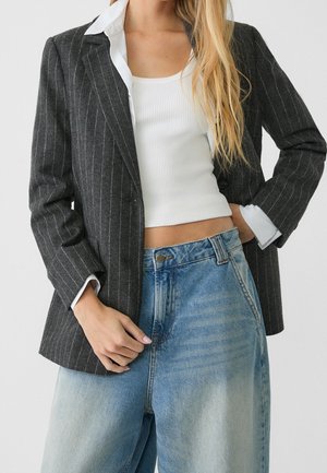 Vrouw draagt een zwarte blazer met krijtstreepjes over een witte geribbelde crop top en loszittende lichtblauwe spijkerbroek, met haar hand rustend op haar heup en de jas.