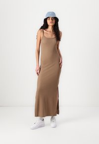 Brązowa maxi sukienka z ribbed, z cienkimi ramiączkami i bocznymi rozcięciami, połączona z białymi sneakersami i jasnoniebieską kapeluszem typu bucket.