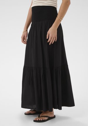 Femme portant une jupe maxi noire à volants avec une taille smockée, un haut rayé, des bracelets en or, des bagues et des sandales noires type tongs.