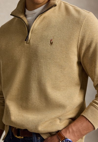 Homme portant un pull beige à fermeture éclair quart, avec un petit logo brodé de cheval, un jean bleu, et une montre en cuir marron avec un cadran bleu.