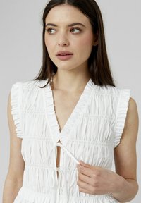 Robe blanche avec des volants verticaux, décolleté en V, manches courtes froncées et fermeture à liens sur le devant. Tissu lisse et texturé avec des fronces.