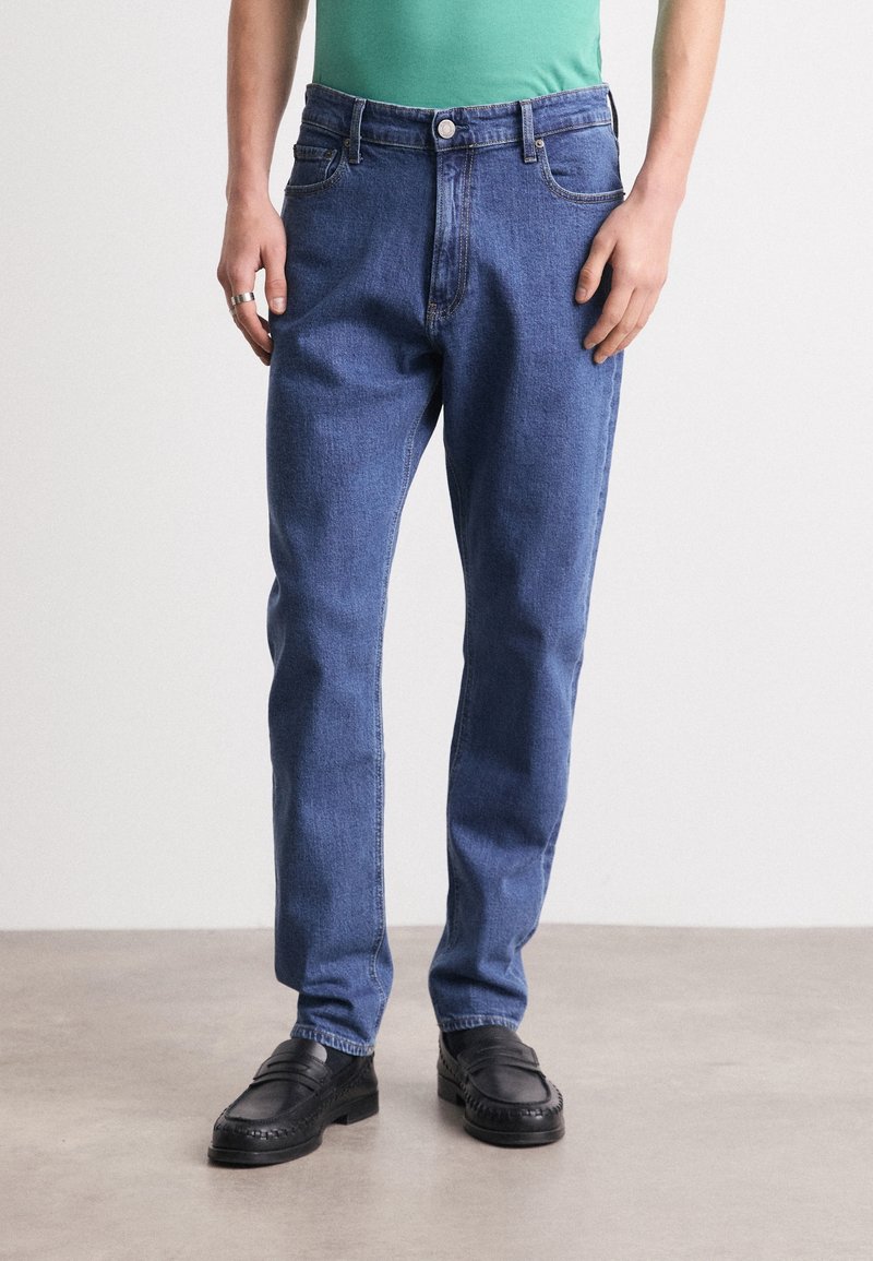 Blå denimjeans med rak benform, fem fickor och knappstängning. Kombinerade med svarta läderskor. Slät textur.