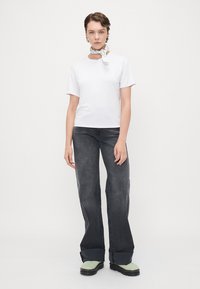 T-shirt bianco a maniche corte, abbinato a jeans grigi a vita alta e svasati. Il modello indossa una sciarpa fantasia e scarpe verdi. Tessuto dalla texture liscia.