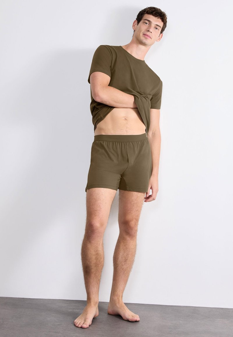Homme portant un t-shirt et un short vert olive, soulevant son t-shirt pour dévoiler son abdomen nu, debout pieds nus contre un mur blanc.