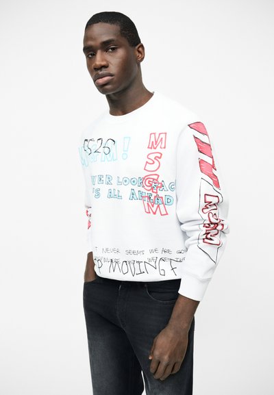 Άνδρας που φοράει λευκό φούτερ με κόκκινο και μπλε γραφικό κείμενο, συνδυασμένο με μαύρο τζιν, στέκεται μπροστά σε λευκό απλό φόντο.