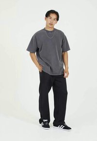 Νέος άνδρας που φοράει χαλαρή γκρι μπλούζα, μαύρες παντελόνες φαρδιές, μαύρα και άσπρα αθλητικά παπούτσια και μια λεπτή ασημένια αλυσίδα, στέκεται με το ένα χέρι στην τσέπη.